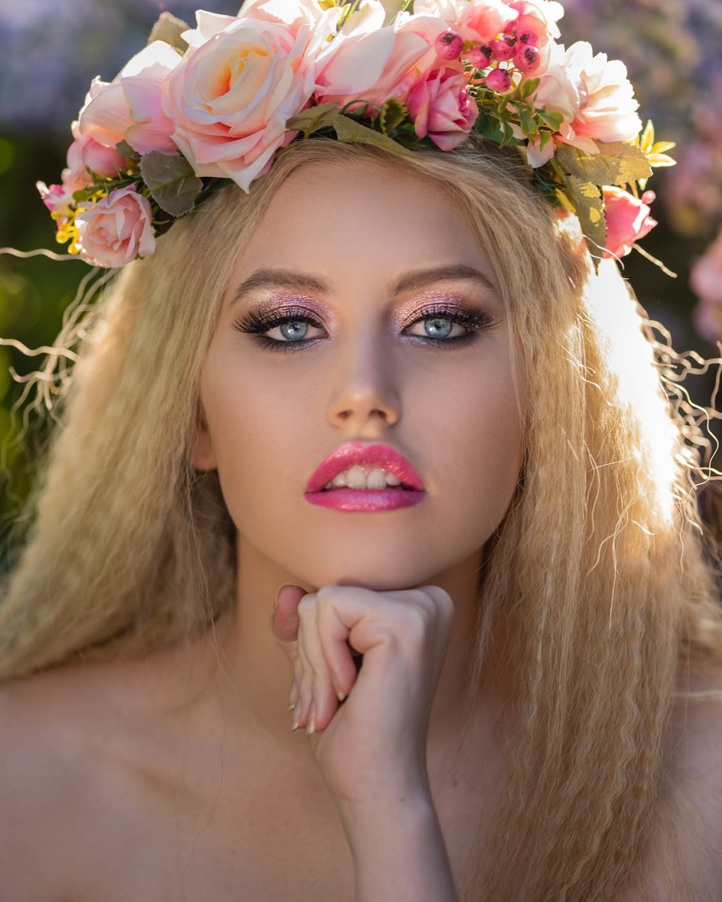 Portret młodej kobiety z blond włosami w wianku z różowych kwiatów, z intensywnym makijażem oczu i ust, opierającej podbródek na dłoni, w miękkim, rozproszonym świetle.