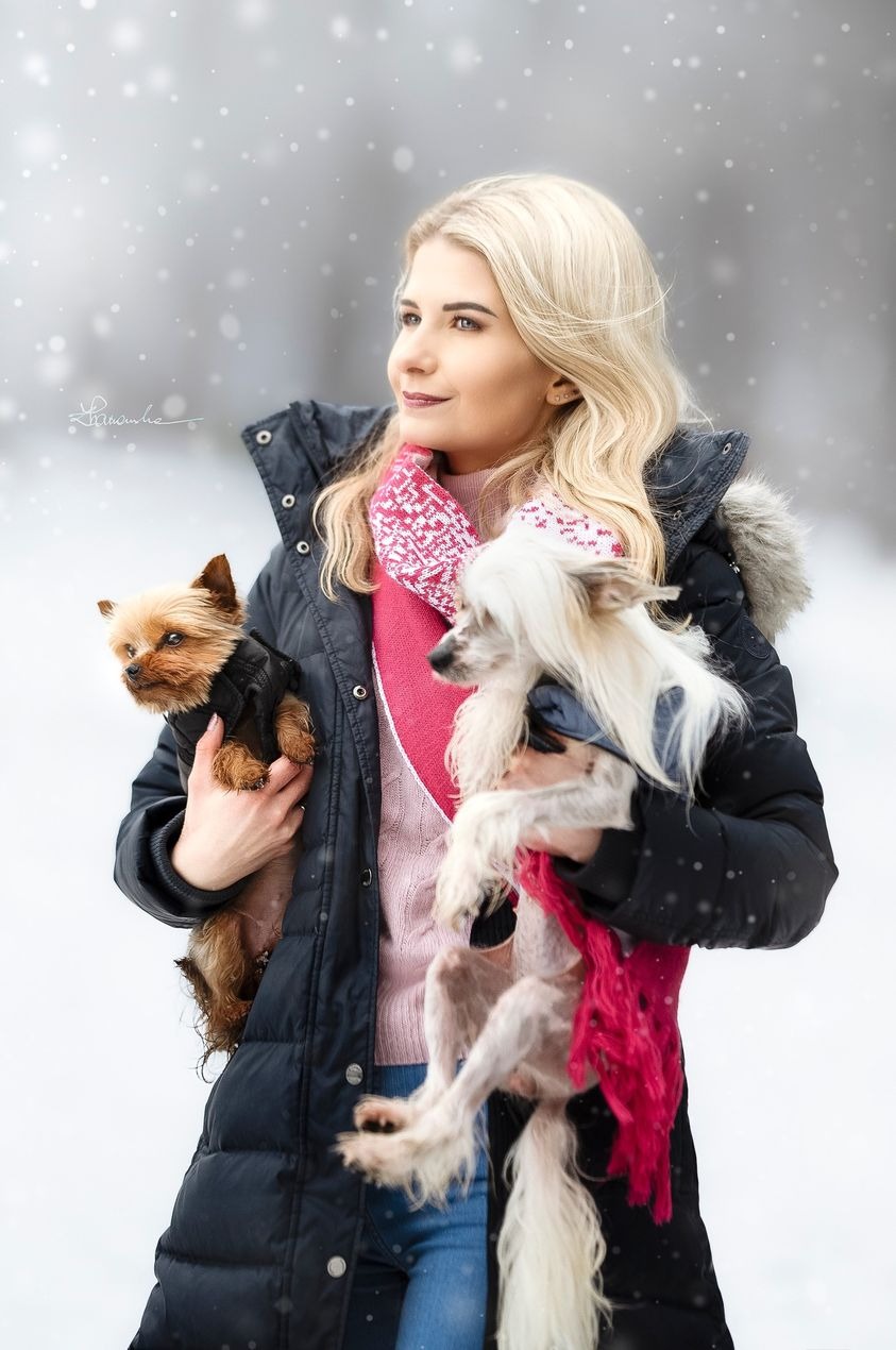 Portret kobiety z blond włosami, ubranej w zimową kurtkę i różowy sweter, trzymającej dwa małe psy na tle zimowego krajobrazu z padającym śniegiem.