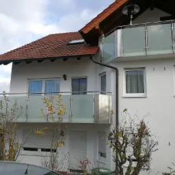 Dwupiętrowy dom z balkonami zabezpieczonymi szklanymi panelami i metalowymi poręczami, widok z dołu na tle pochmurnego nieba.