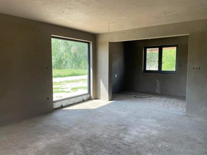 Surowe wnętrze po nałożeniu tynków: widoczne ściany i podłoga w szarym kolorze, duże okno z widokiem na zieleń na zewnątrz, małe okno z widoczną drabiną na zewnątrz, przygotowane do dalszych prac...