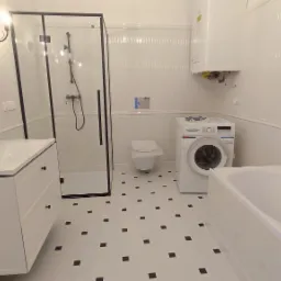 Biała łazienka po remoncie z czarnymi akcentami: kabina prysznicowa z czarną ramą, umywalka z szafką, bateria w stylu retro, pralka, bojler, toaleta w tle i podłoga w biało-czarną szachownicę.