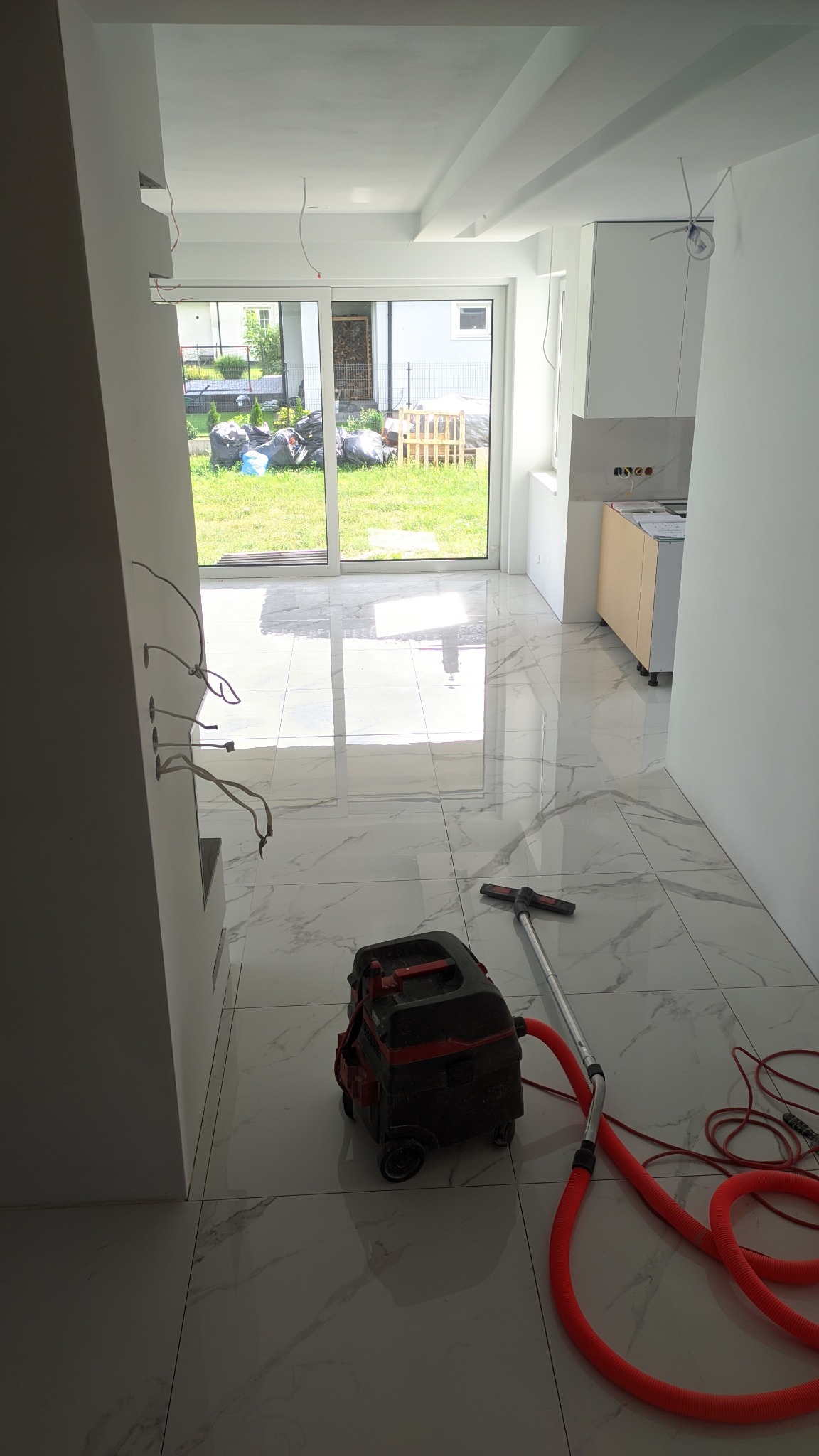 Błyszcząca, marmurowa podłoga z odkurzaczem przemysłowym w nowym, jasnym wnętrzu z widokiem na ogród przez przesuwne drzwi balkonowe.