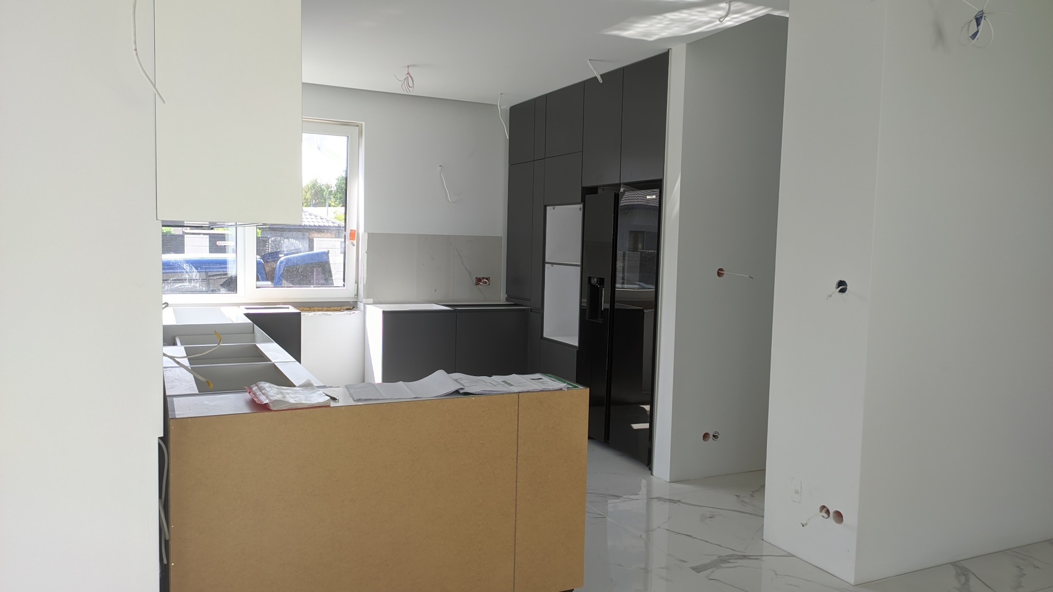 Wnętrze kuchni w trakcie prac wykończeniowych: widoczne elementy zabudowy kuchennej w odcieniach szarości i bieli, niezabudowana wyspa kuchenna z MDF, marmurowe płytki podłogowe oraz wystające...