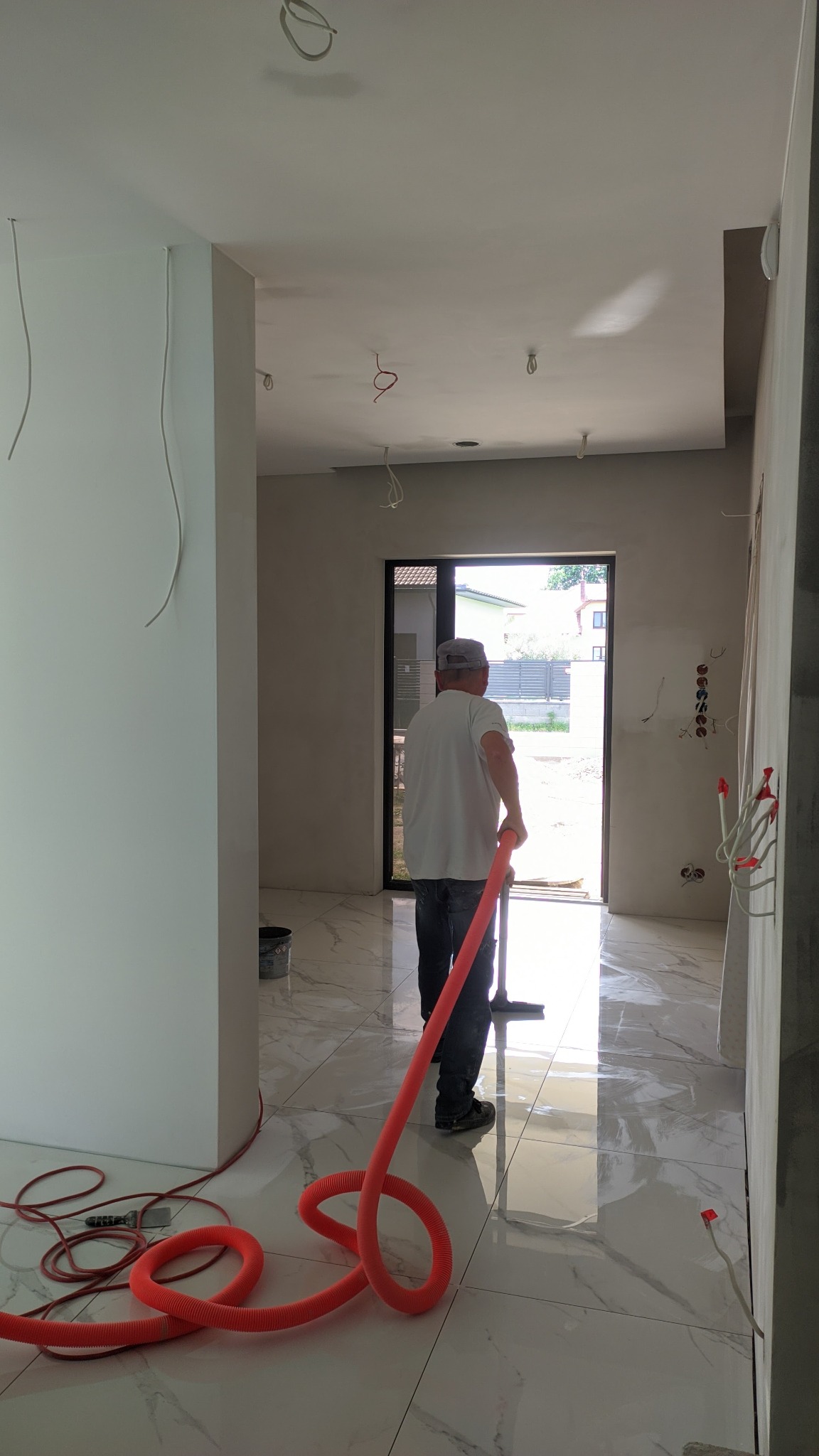 Mężczyzna w czapce sprząta podłogę z płytek imitujących marmur, używając odkurzacza przemysłowego z pomarańczowym wężem; widoczne niedokończone ściany i instalacje elektryczne.