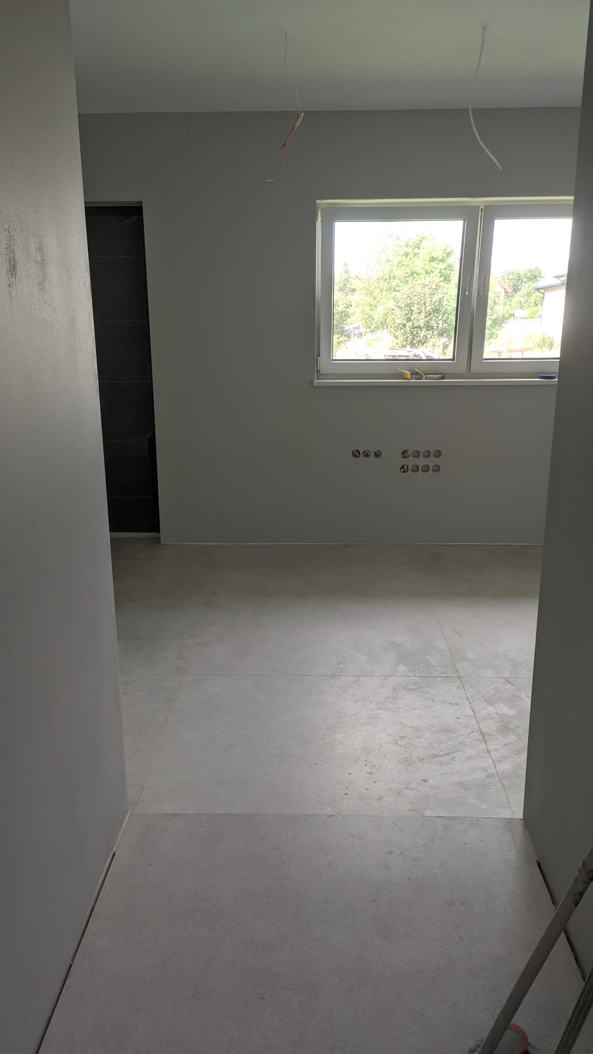 Surowe wnętrze z szarymi ścianami i podłogą w trakcie wykańczania, widoczne wystające kable elektryczne i otwory pod gniazdka, okno z widokiem na zieleń, częściowo widoczna drabina malarska...
