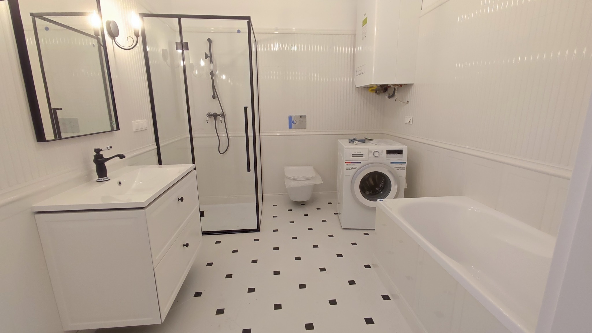 Biała łazienka po remoncie z czarnymi akcentami: kabina prysznicowa z czarną ramą, umywalka z szafką, bateria w stylu retro, pralka, bojler, toaleta w tle i podłoga w biało-czarną szachownicę.