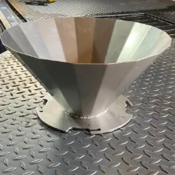 Stożkowy element metalowy z podstawą, wykonany z blachy i spawany, leżący na metalowej powierzchni z fakturą łezki.