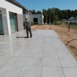 Pracownik myje wodą pod ciśnieniem nowo ułożony taras z szarych płyt betonowych na zewnątrz nowoczesnego domu.