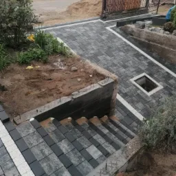 Kaskadowe schody i chodnik z szaro-białej kostki brukowej, w trakcie budowy, z widoczną ziemią i materiałami budowlanymi, z metalowymi prętami wystającymi z betonu.