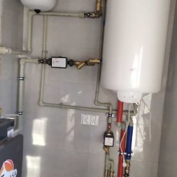 Konrad Wardak - Wnętrze kotłowni z białym zbiornikiem wyrównawczym, bojlerem i licznymi rurami z zaworami i filtrami, widoczne gniazdka elektryczne na ścianie z jasnych płytek.