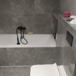 Nowoczesna łazienka z wanną obudowaną szarymi płytkami imitującymi marmur, czarną armaturą i elementami wyposażenia, widok z perspektywy użytkownika.