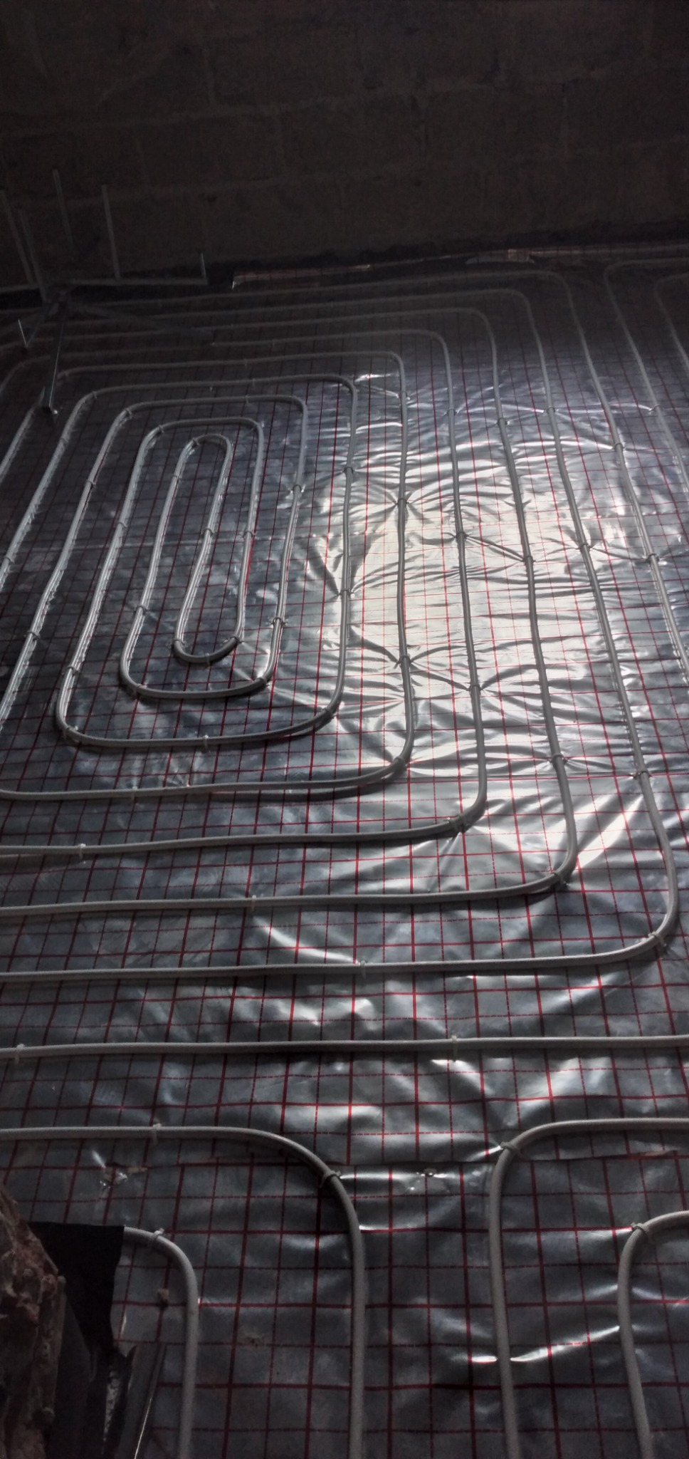 Rozłożone rury ogrzewania podłogowego na folii z siatką w pomieszczeniu w trakcie budowy, widoczne mocowania rur.