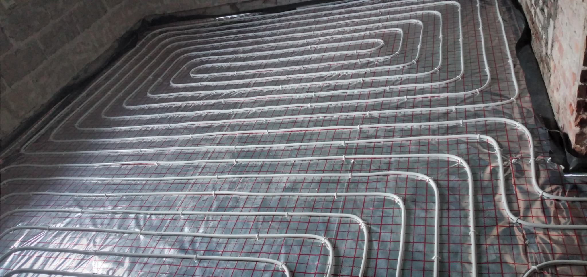 Rozłożone białe rury ogrzewania podłogowego na folii z czerwoną siatką, widoczne surowe ściany w tle.