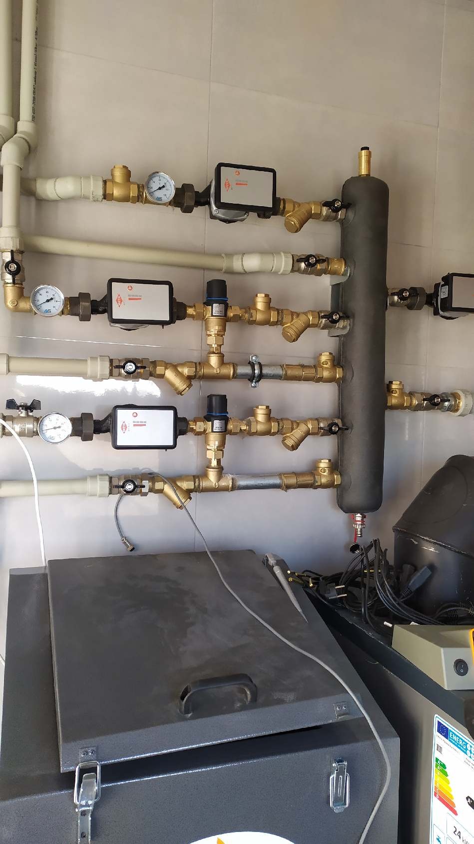 Rozbudowana instalacja hydrauliczna z licznymi zaworami, manometrami i czujnikami zamontowana na jasnej ścianie, z widocznym szarym zbiornikiem w dolnej części kadru.