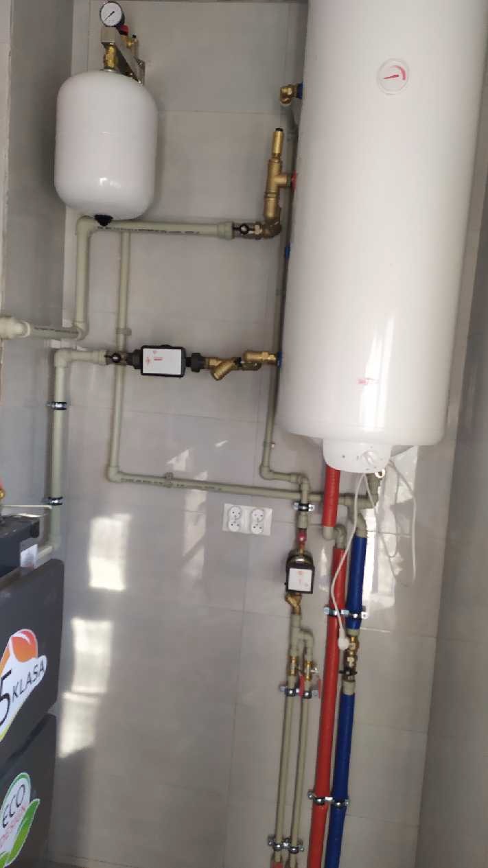 Wnętrze kotłowni z białym zbiornikiem wyrównawczym, bojlerem i licznymi rurami z zaworami i filtrami, widoczne gniazdka elektryczne na ścianie z jasnych płytek.