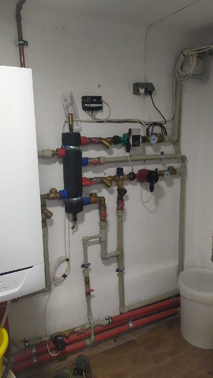 Węzeł cieplny z kotłem gazowym, pompami obiegowymi, zaworami i orurowaniem, zamontowany na ścianie w pomieszczeniu technicznym. Widoczne przewody elektryczne i elementy sterujące.