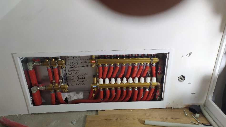 Rozdzielacz ogrzewania podłogowego z czerwonymi rurami, zamontowany we wnęce w ścianie, z notatkami na ścianie obok.