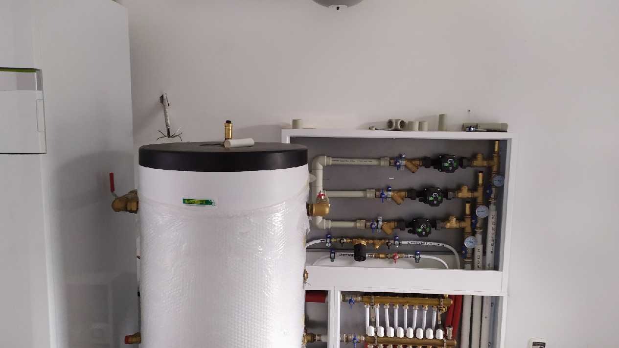 Biały zasobnik ciepłej wody owinięty folią bąbelkową, obok rozdzielacz podłogowy i instalacja grzewcza z rurami, zaworami i manometrami w szafce w pomieszczeniu technicznym.