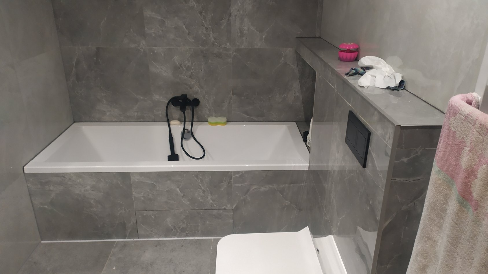 Nowoczesna łazienka z wanną obudowaną szarymi płytkami imitującymi marmur, czarną armaturą i elementami wyposażenia, widok z perspektywy użytkownika.