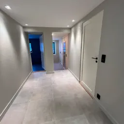 Korytarz z szarymi, kwadratowymi płytkami podłogowymi, białymi drzwiami z czarnymi klamkami i oświetleniem punktowym w suficie.