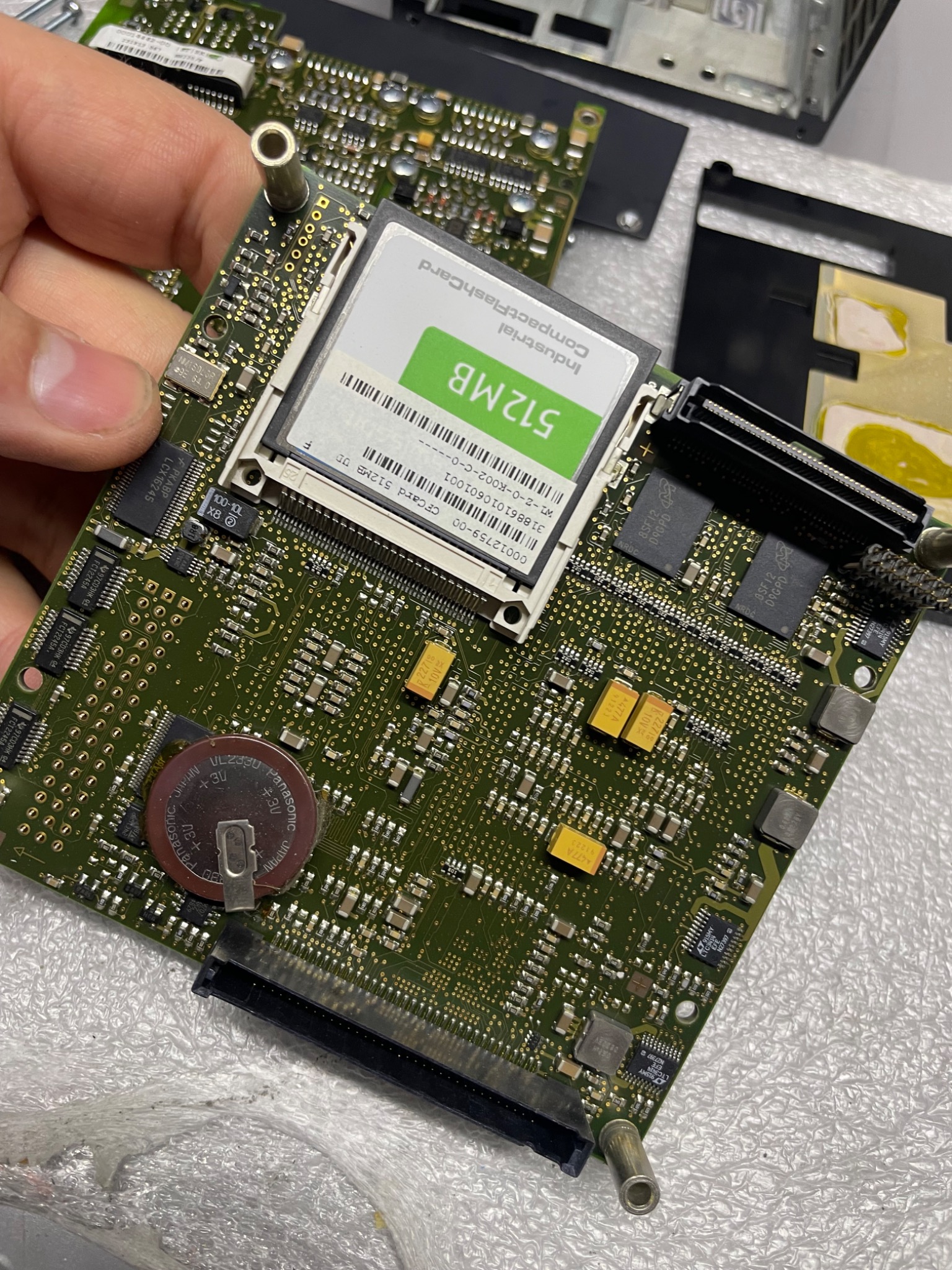 Szczegółowe zbliżenie na zieloną płytę główną komputera z widocznymi elementami elektronicznymi, w tym baterią pastylkową i kartą CompactFlash 512MB, trzymaną w dłoni.