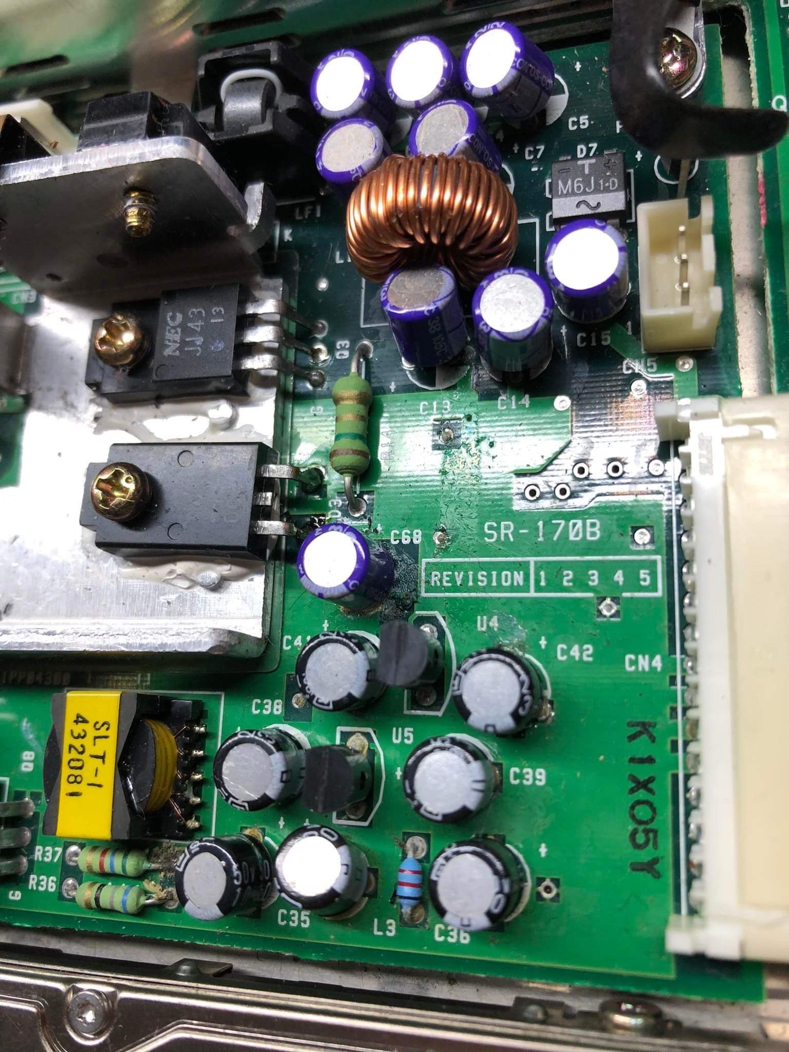 Zbliżenie na zieloną płytę główną z elementami elektronicznymi, w tym kondensatory, rezystory, transformator toroidalny z miedzianym uzwojeniem, oraz układ scalony NEC JJ43, widoczne oznaczenia...