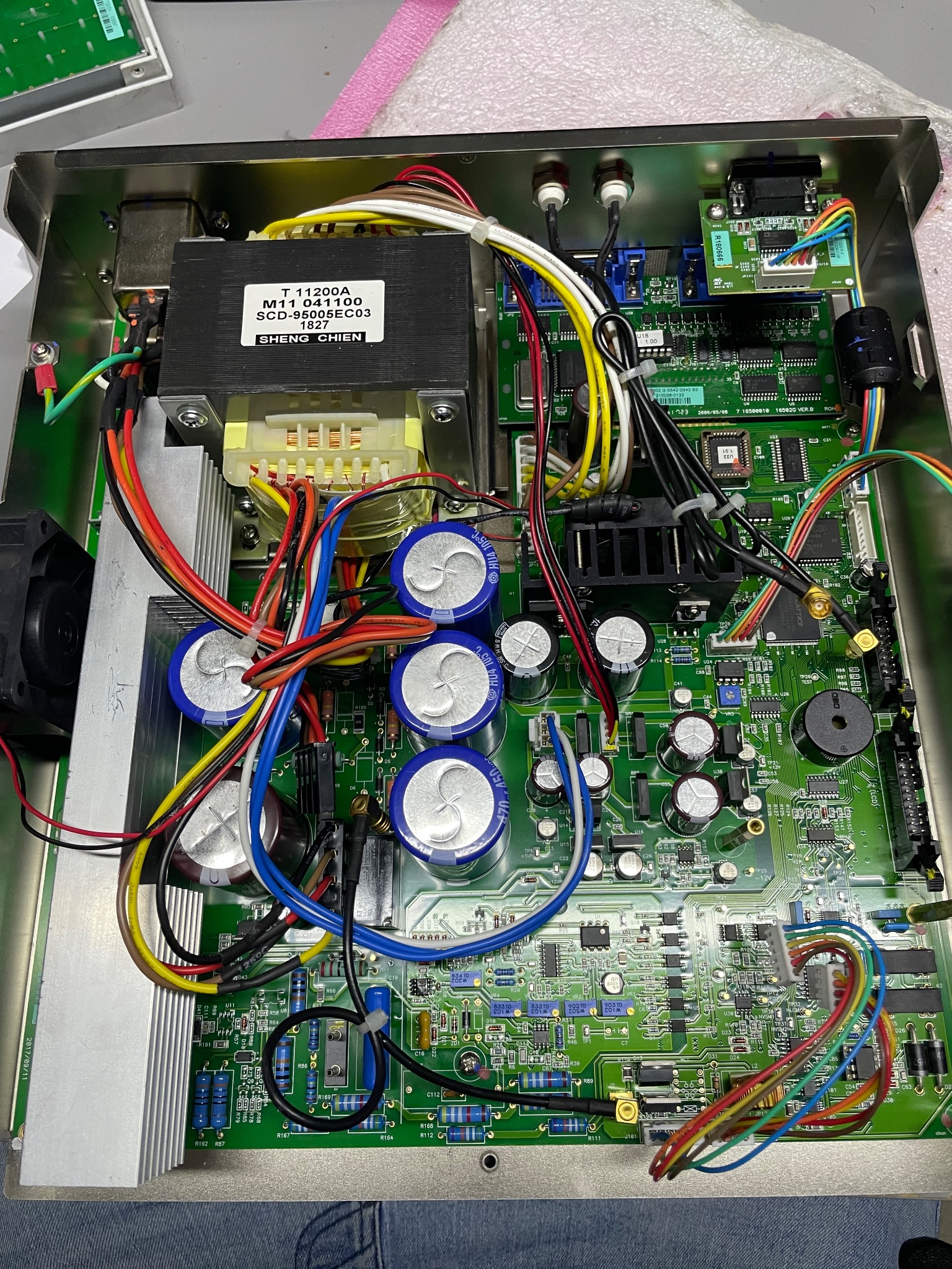 Otwarta obudowa urządzenia elektronicznego z widoczną zieloną płytą główną, kondensatorami, transformatorem i licznymi kablami w różnych kolorach.