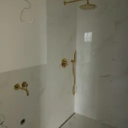 Nowoczesna kabina prysznicowa z białymi płytkami imitującymi marmur i złotą armaturą, widoczne wystające kable elektryczne w górnym rogu.