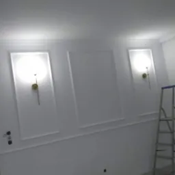 Białe ściany z dekoracyjnymi panelami i zamontowanymi kinkietami, widoczna drabina malarska.