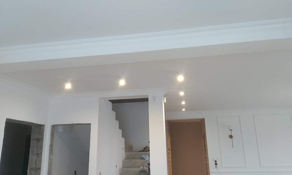 Białe wnętrze w trakcie remontu z widocznymi schodami, sufit z listwami dekoracyjnymi i oświetleniem punktowym, ściany z dekoracyjnymi panelami.