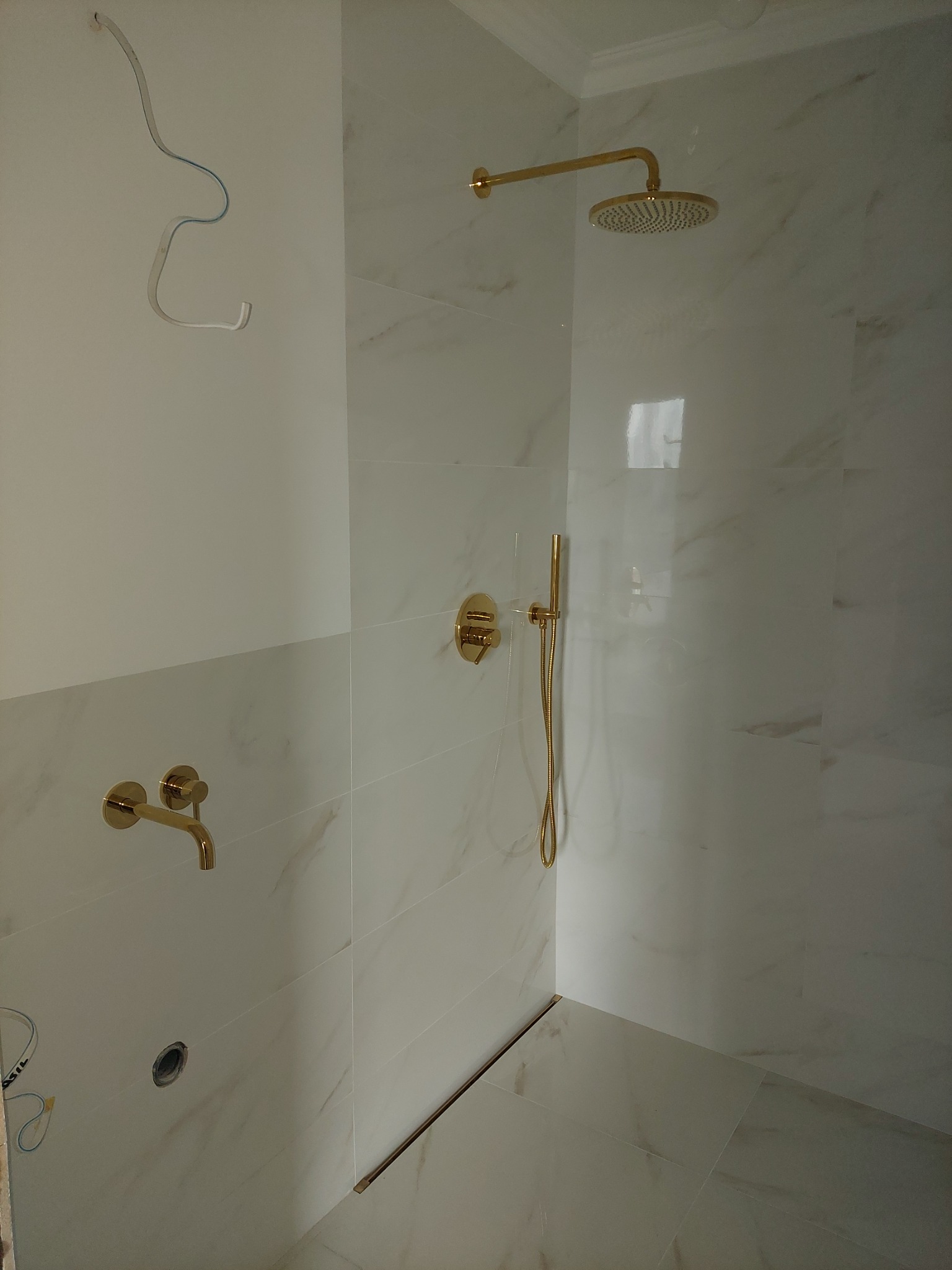 Nowoczesna kabina prysznicowa z białymi płytkami imitującymi marmur i złotą armaturą, widoczne wystające kable elektryczne w górnym rogu.