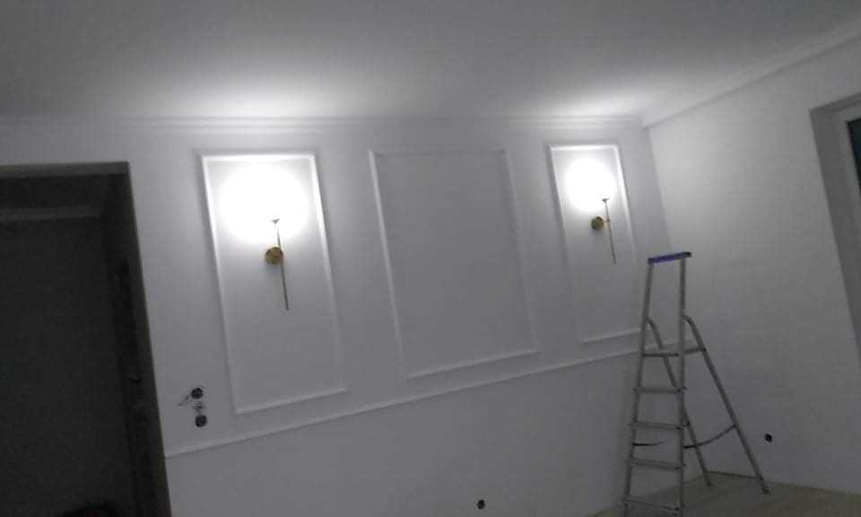 Białe ściany z dekoracyjnymi panelami i zamontowanymi kinkietami, widoczna drabina malarska.