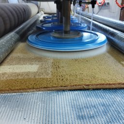 Multisystem Jarosław Stosio - Automatyczna maszyna z obrotowymi, niebieskimi szczotkami czyści duży, brązowo-beżowy dywan w procesie produkcyjnym, widoczne rolki prowadzące i system natrysku wody.