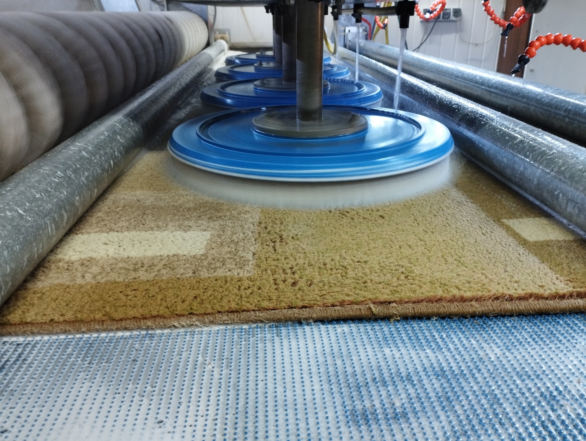 Automatyczna maszyna z obrotowymi, niebieskimi szczotkami czyści duży, brązowo-beżowy dywan w procesie produkcyjnym, widoczne rolki prowadzące i system natrysku wody.