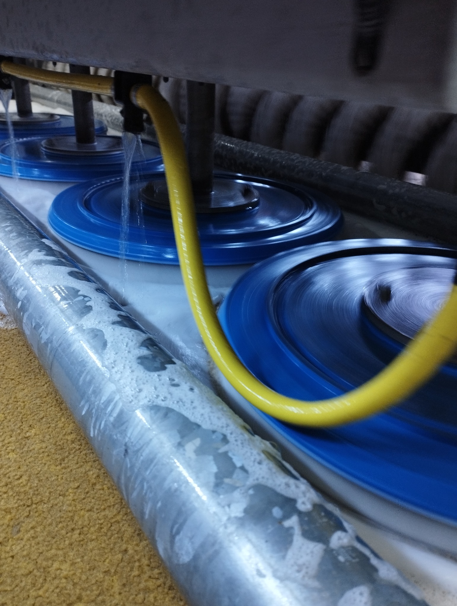 Automatyczne urządzenie z rotującymi, niebieskimi szczotkami i żółtym wężem doprowadzającym wodę, czyszczące beżowy dywan, widoczne ślady piany.