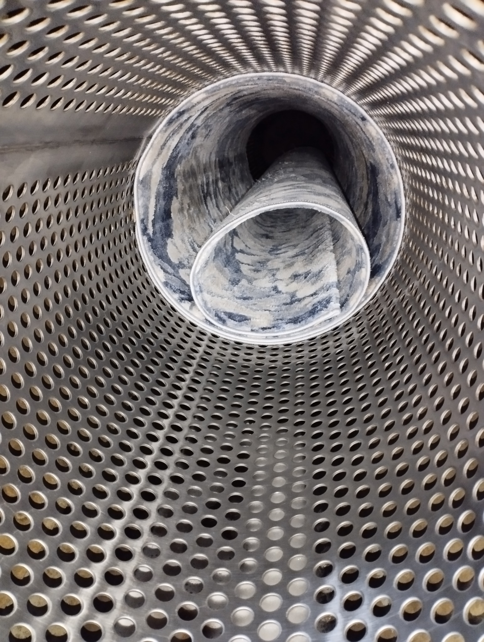 Wnętrze perforowanego bębna pralki przemysłowej z widocznym zwiniętym dywanem w środku.