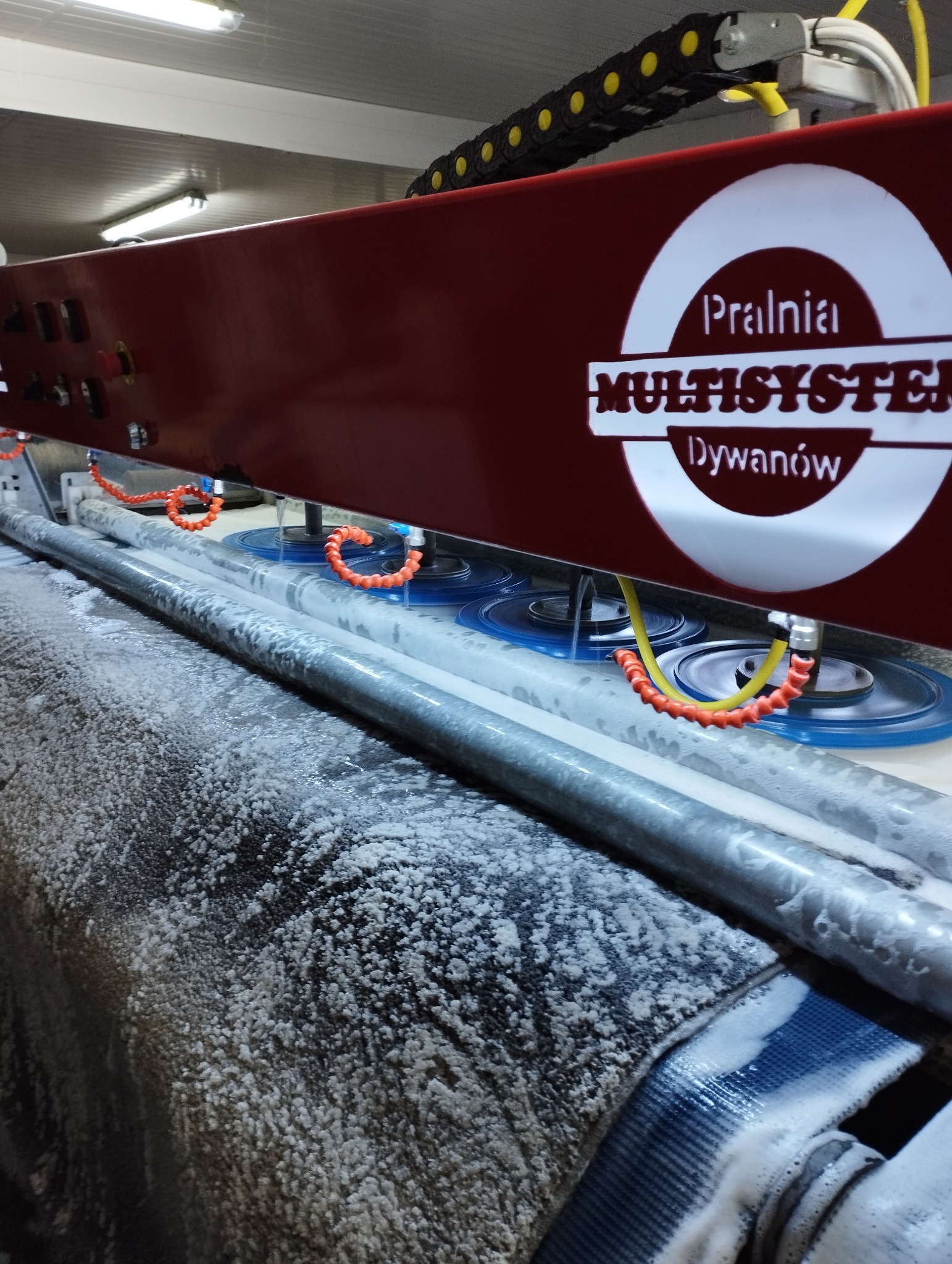 Automatyczna maszyna z logiem 'Pralnia Multisystem Dywanów' w trakcie czyszczenia dywanu, widoczne obracające się szczotki i piana.