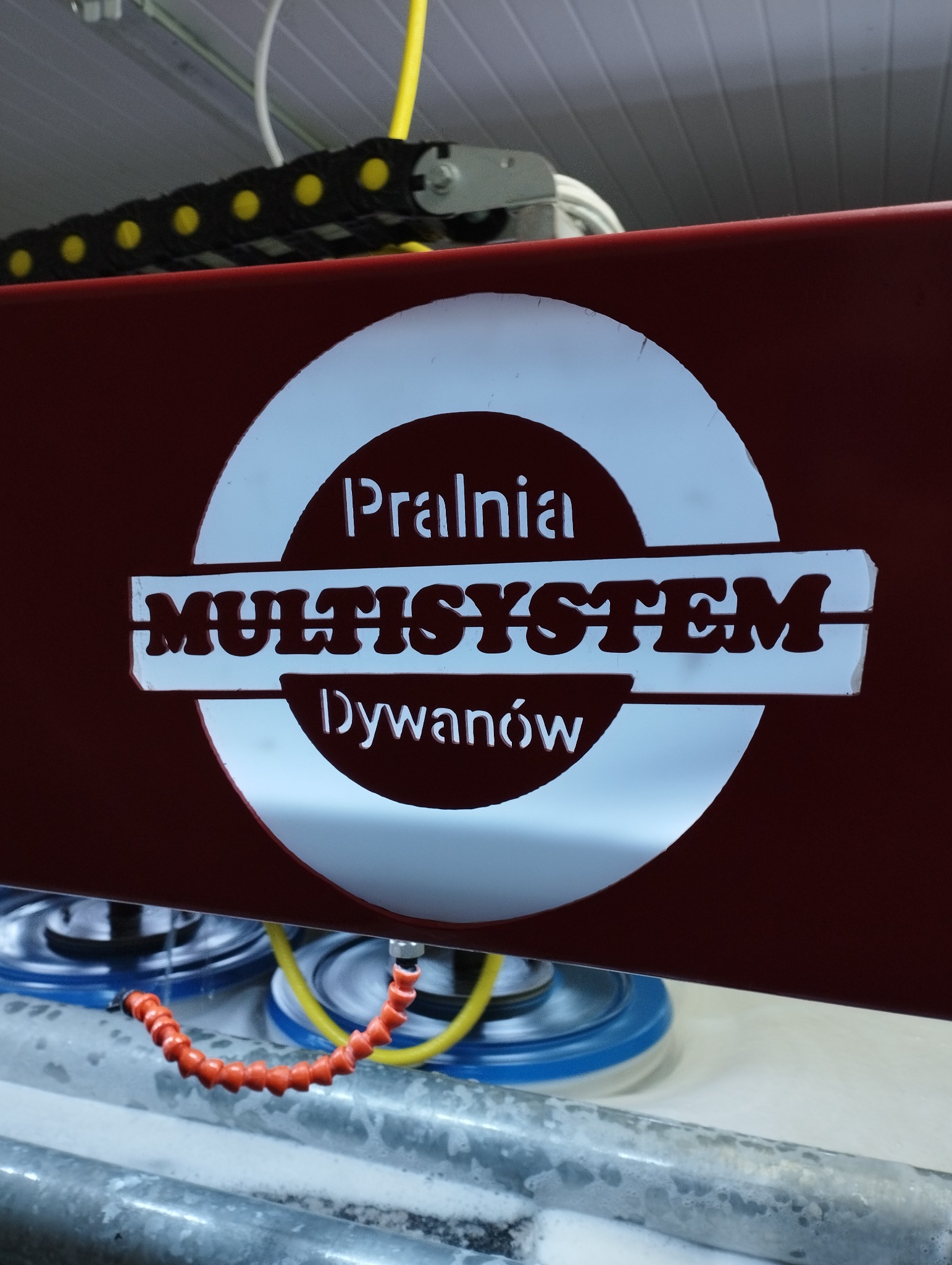 Czerwony panel maszyny z logo 'Pralnia Multisystem Dywanów' nad wirującymi szczotkami i pianą, widok z bliska.
