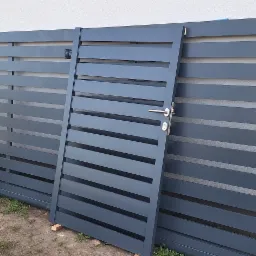 Elementy metalowego ogrodzenia z poziomymi panelami i furtką z klamką, oparte o ścianę budynku w trakcie montażu.