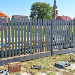 Nowoczesne, szare ogrodzenie panelowe z przesuwną bramą wjazdową, widoczne narzędzia i materiały budowlane na trawie, w tle dom w budowie i kościół.