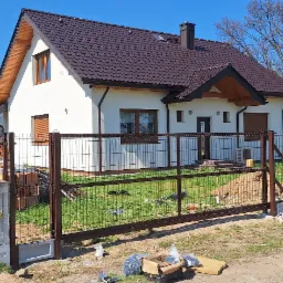 Brązowe ogrodzenie panelowe z bramą przesuwną przed białym domem z brązowym dachem, widoczna skrzynka elektryczna, narzędzia i materiały montażowe na trawniku.