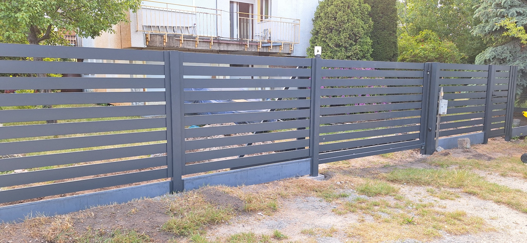 Nowoczesne szare ogrodzenie panelowe z poziomymi lamelami, widok z zewnątrz posesji, zarośnięty teren wokół ogrodzenia.