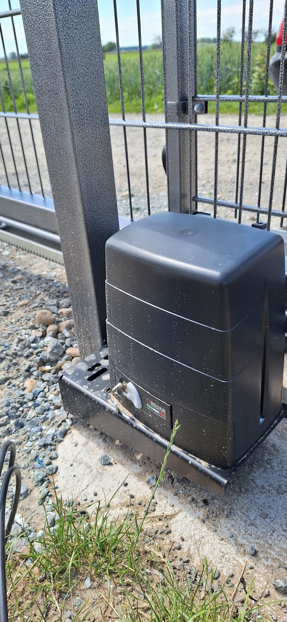 Czarny automat do bram przesuwnych marki Roger, zamontowany przy metalowej konstrukcji, z widoczną zębatką i fragmentem ogrodzenia panelowego w tle.