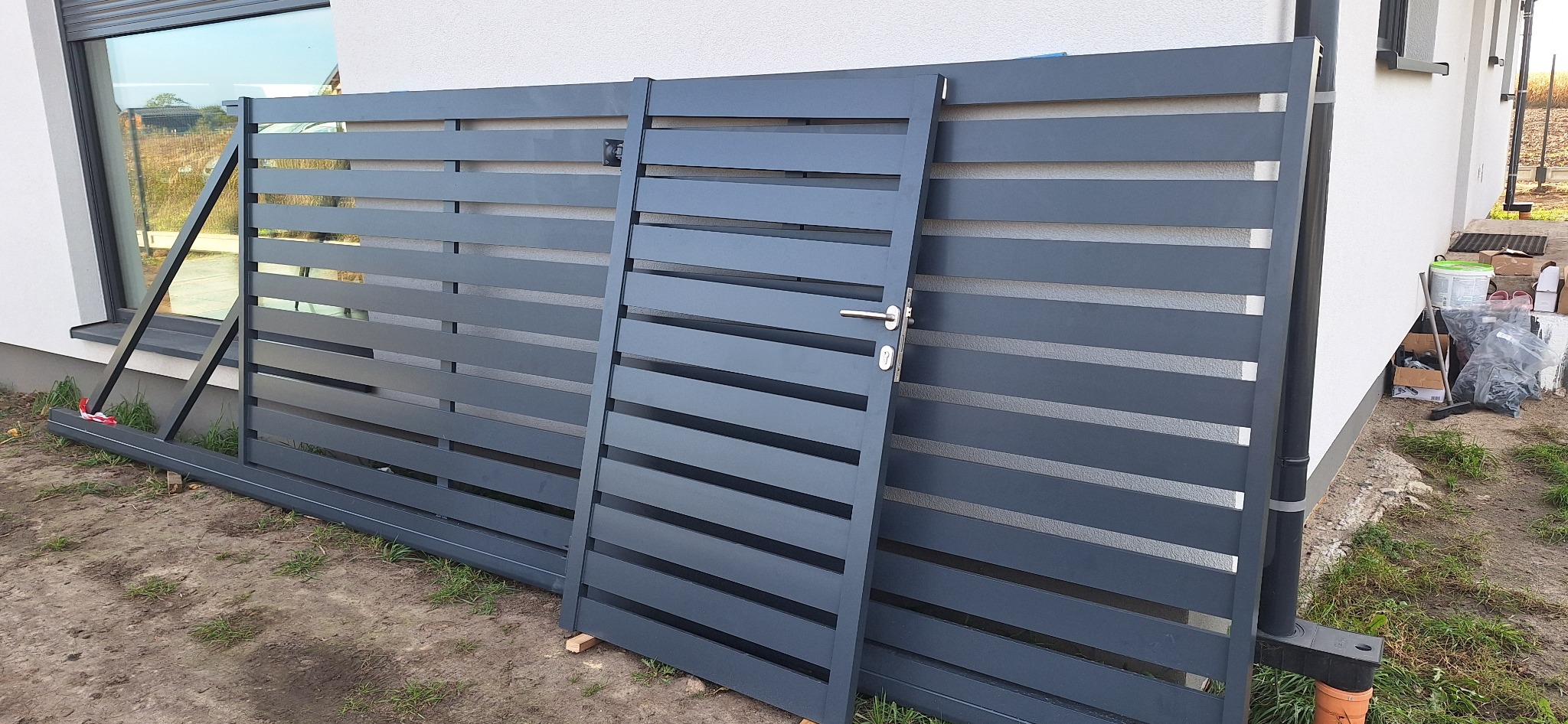 Elementy metalowego ogrodzenia z poziomymi panelami i furtką z klamką, oparte o ścianę budynku w trakcie montażu.