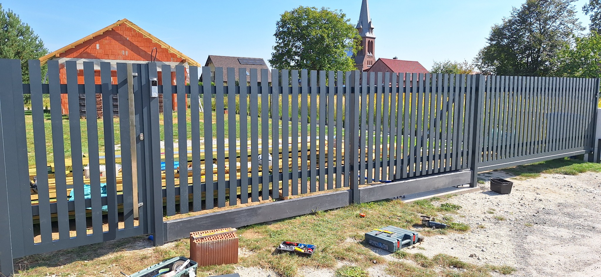 Nowoczesne, szare ogrodzenie panelowe z przesuwną bramą wjazdową, widoczne narzędzia i materiały budowlane na trawie, w tle dom w budowie i kościół.