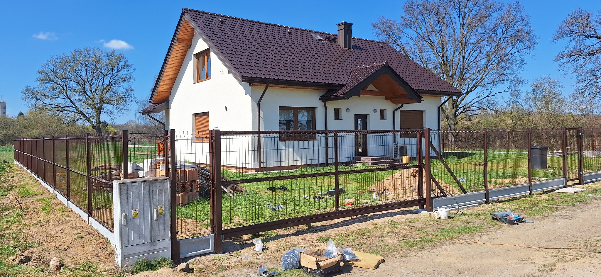 Brązowe ogrodzenie panelowe z bramą przesuwną przed białym domem z brązowym dachem, widoczna skrzynka elektryczna, narzędzia i materiały montażowe na trawniku.