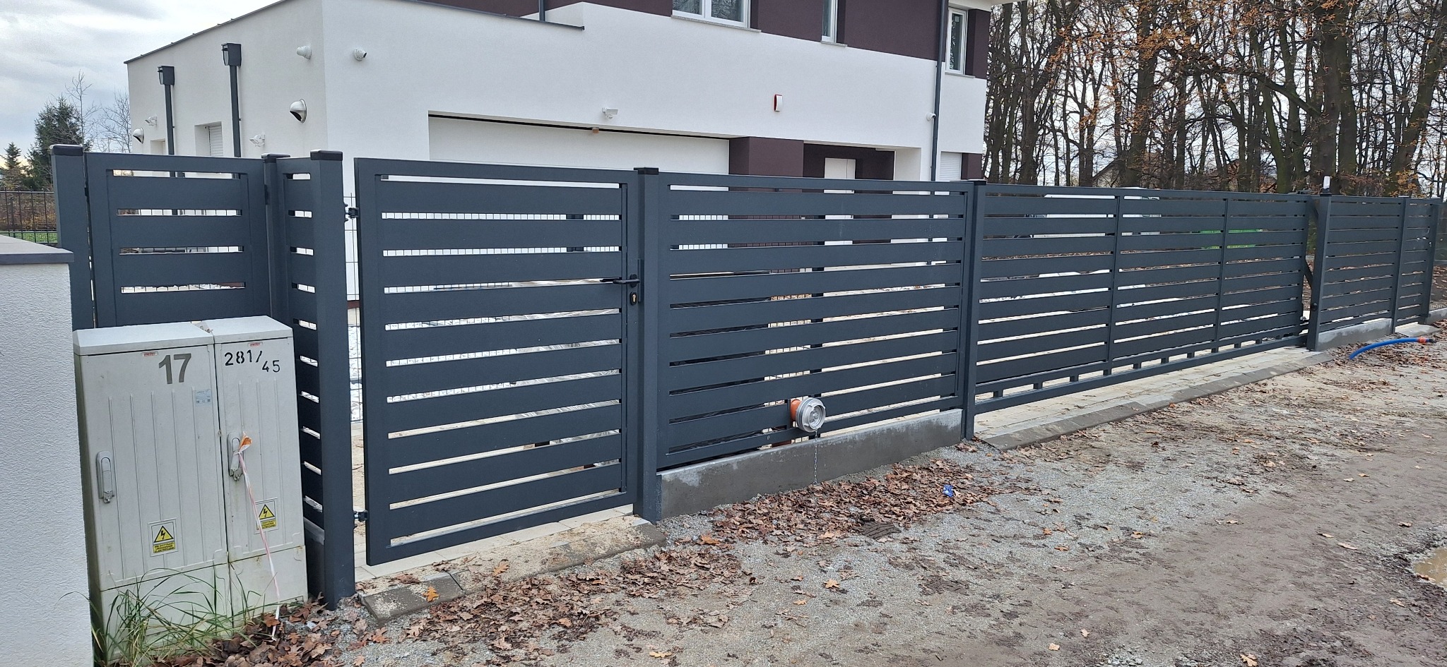 Nowoczesne, poziome ogrodzenie metalowe w kolorze grafitowym z bramą wjazdową, na tle białego domu z szarymi akcentami. Widoczne skrzynki elektryczne.
