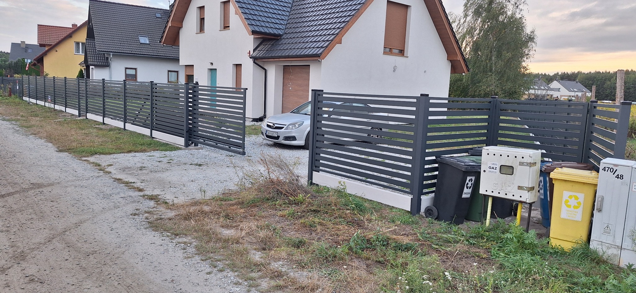 Nowoczesne, poziome ogrodzenie metalowe w kolorze grafitowym, z otwartą bramą wjazdową na posesję z białym domem i zaparkowanym samochodem. Widoczne kosze na śmieci.