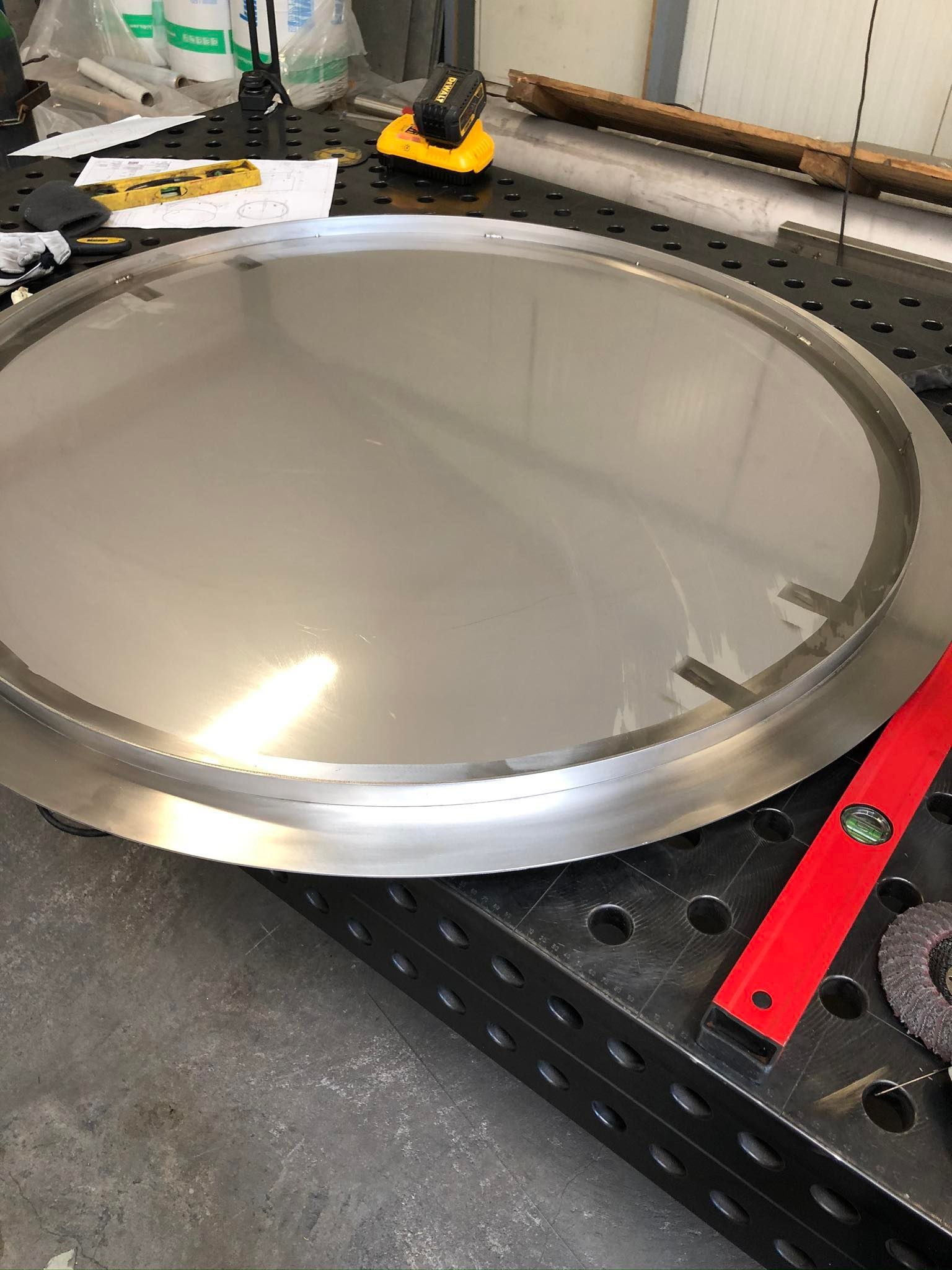 Okrągły element ze stali nierdzewnej z widocznymi śladami obróbki, leżący na perforowanym stole warsztatowym obok poziomicy, szlifierki listkowej i rysunku technicznemu.