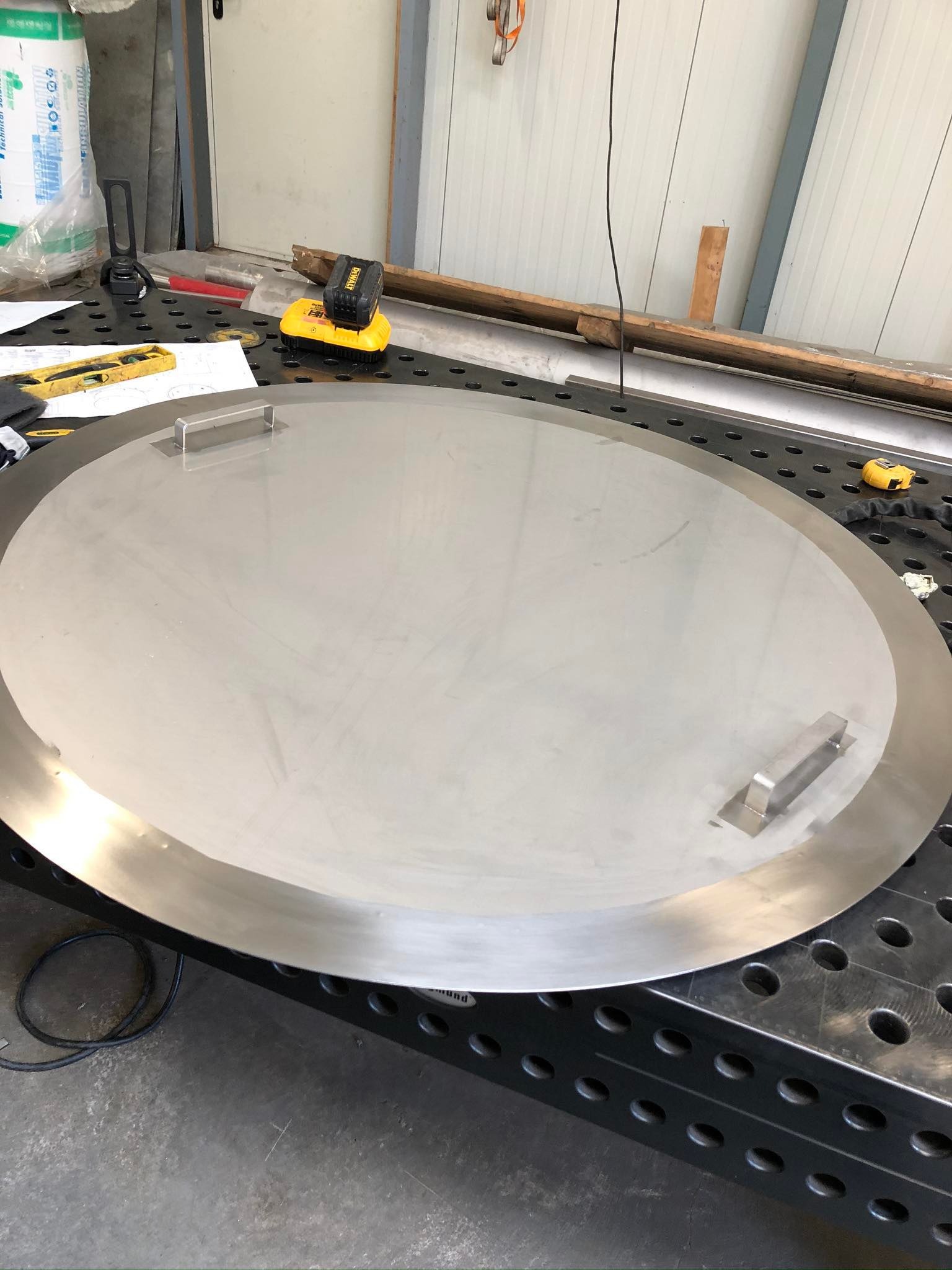 Okrągła pokrywa ze stali nierdzewnej z uchwytami, leżąca na perforowanym stole warsztatowym, obok narzędzi i akumulatora DeWalt.